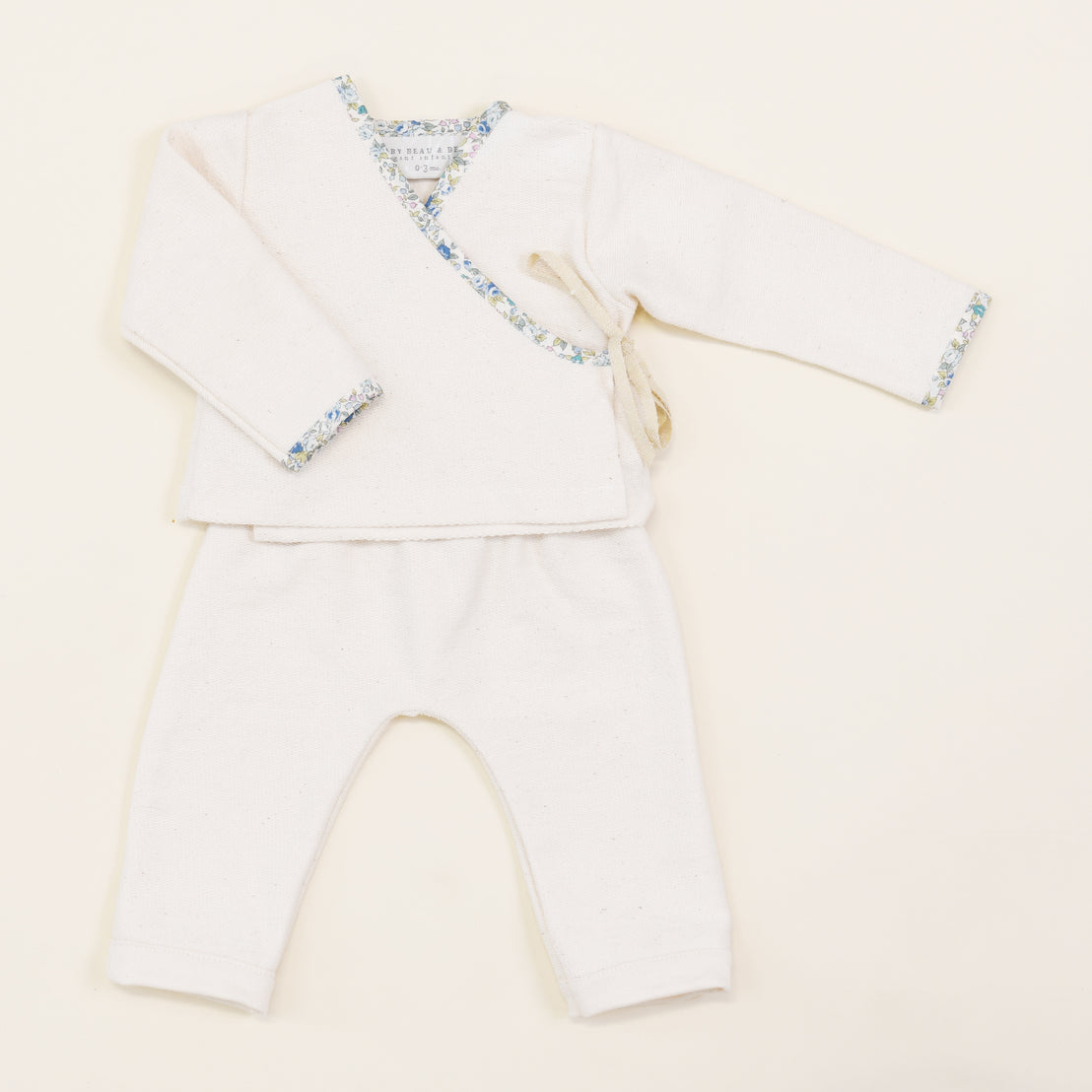 An upscale, boutique Petite Fleur Wrap Top & Pants baby kimono top with floral trim and matching white pants, displayed flat on a light beige background.