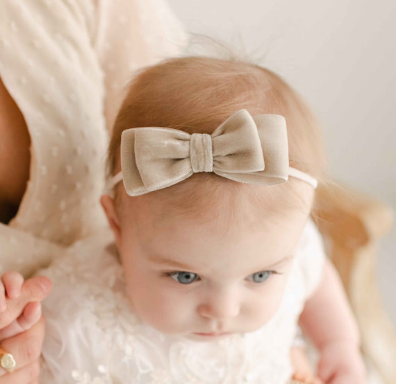 Close-up of a baby with blue eyes wearing a Kristina Velvet Bow Headband, held in the arms of an adult during a christening.