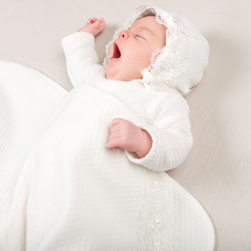 A newborn baby dressed in a white heirloom outfit with lace detailing and a bonnet yawns while wrapped in the Madeline Personalized Blanket.