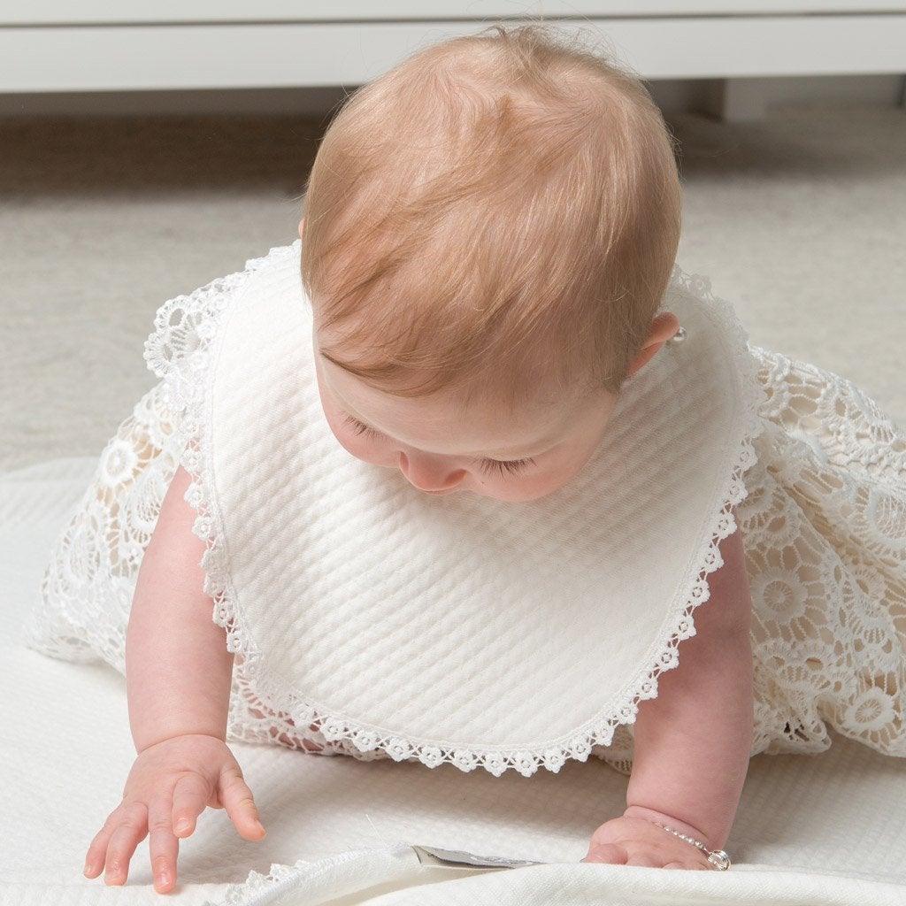 A baby crawls on the ground wearing a christening bib with scalloped edges. 