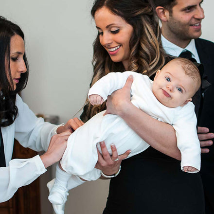 Godparent Gift Etiquette: A godmother helping dress a baby as part of a baptism. Baby is being held by mother with a funny face.