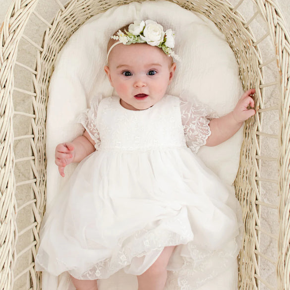 A baby wearing a white dress and a floral headband lies in a white wicker bassinet, looking up with a curious expression.