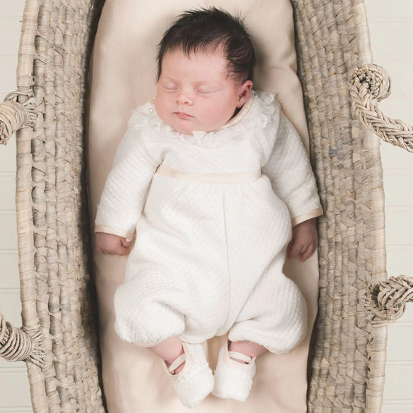 A baby with dark hair, wearing a white outfit and shoes, is peacefully sleeping in a woven basket with handles. The baby rests on a cream-colored cushion inside the basket.