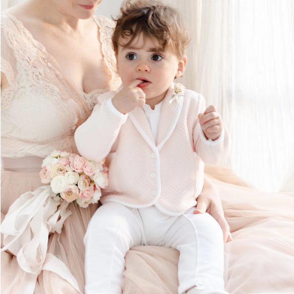 A woman in a beige lace dress holds a baby dressed in a pink jacket and white pants. The baby has curly hair and is touching their lips. The woman holds a small bouquet of light pink and white flowers. Light flows in from the background.