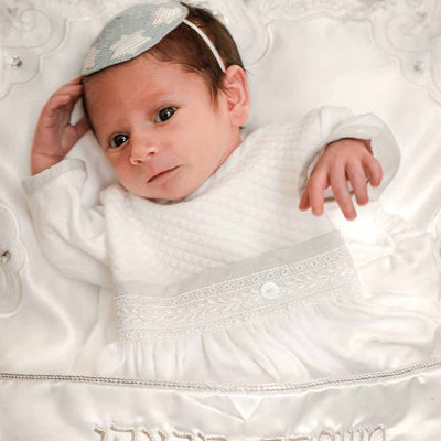 A baby lies on a white satin pillow while wearing a white outfit with delicate lace details and a knitted white and gray kippah. The background is also white, with embroidered patterns. The baby has a calm expression.