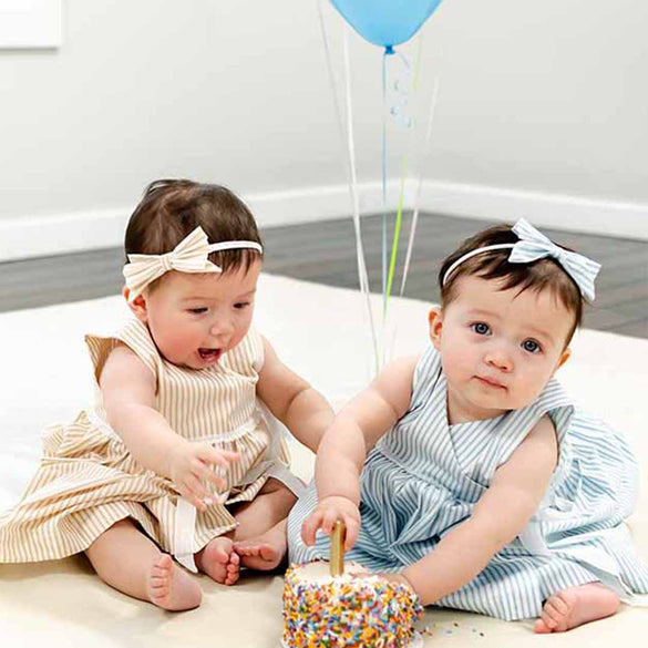 Two babies wearing matching bows and striped dresses are seated on the floor. One baby reaches excitedly toward a sprinkle-covered cake with a candle, while the other baby looks at the camera. Blue balloons are in the background.