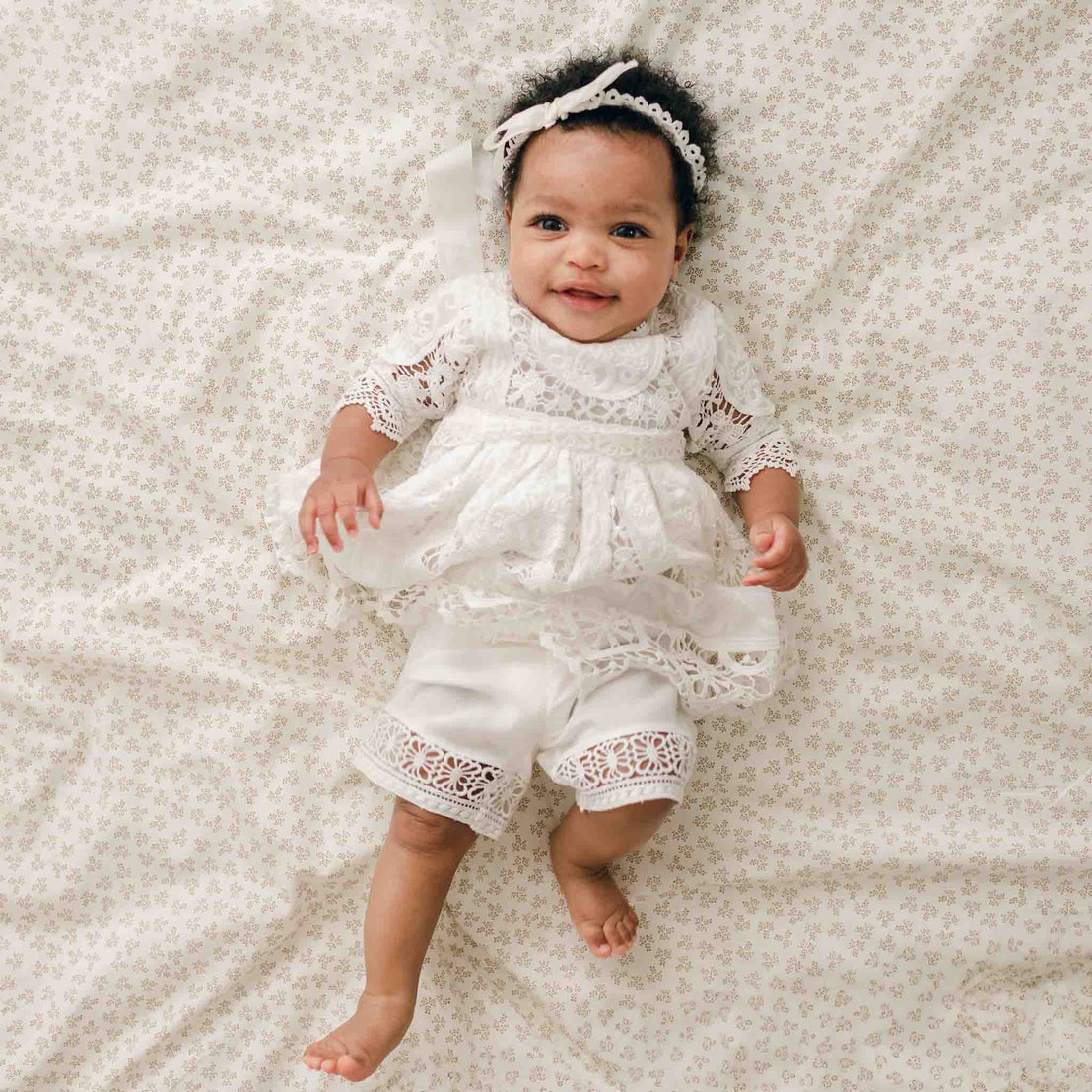 A baby is lying on a floral-patterned blanket, dressed in the stunning Adeline Lace Dress & Bloomers with a matching headband. Their outfit, perfect for any special occasion like a christening, includes a lace-trimmed top with a bow and 100% cotton lace-trimmed bloomers. The baby smiles while looking up.