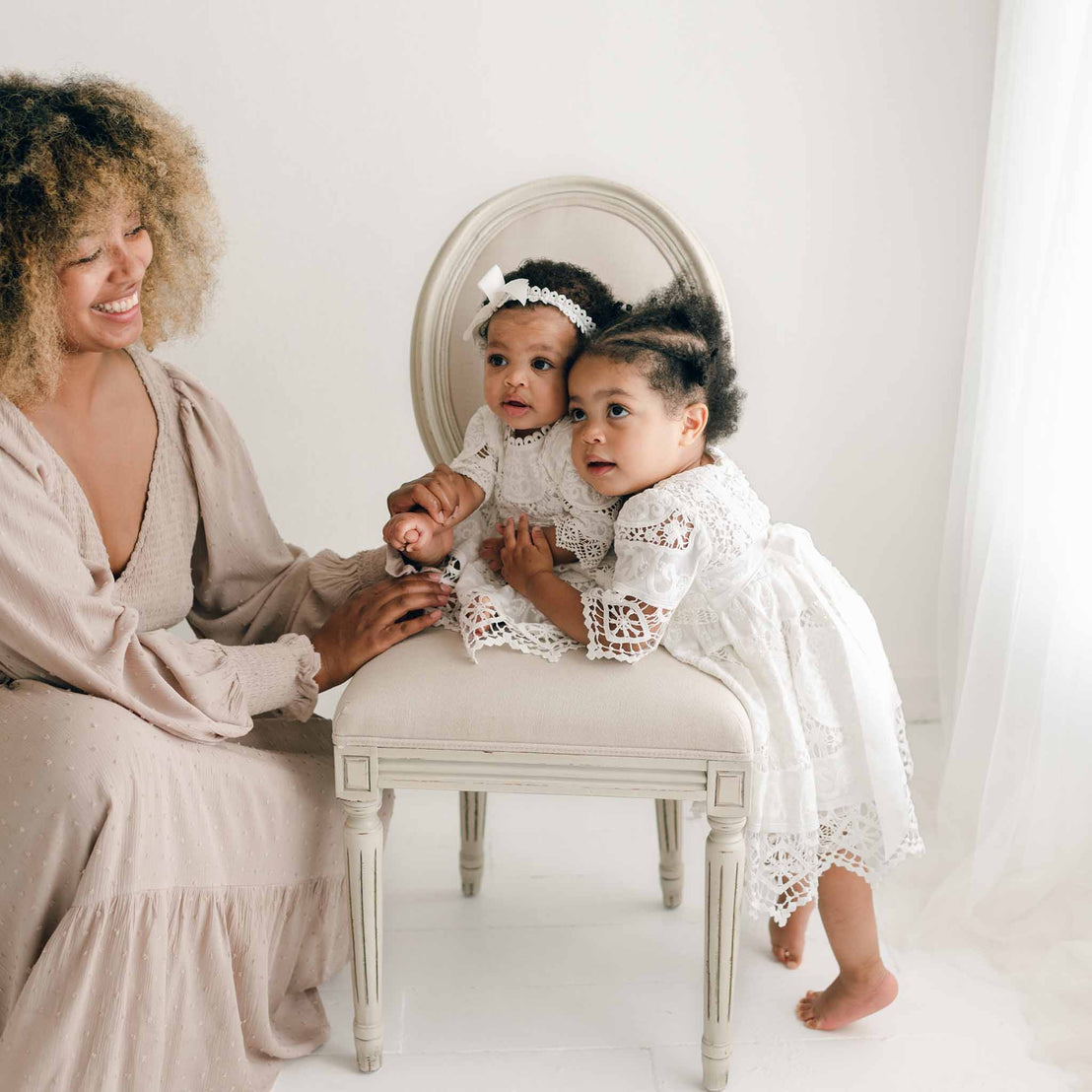 A woman with curly hair smiles at two young children dressed in Adeline Lace Dress & Bloomers. One child sits on a cream-colored chair, while the other stands beside it, looking away. The room features a bright, clean aesthetic with minimalistic white decor.