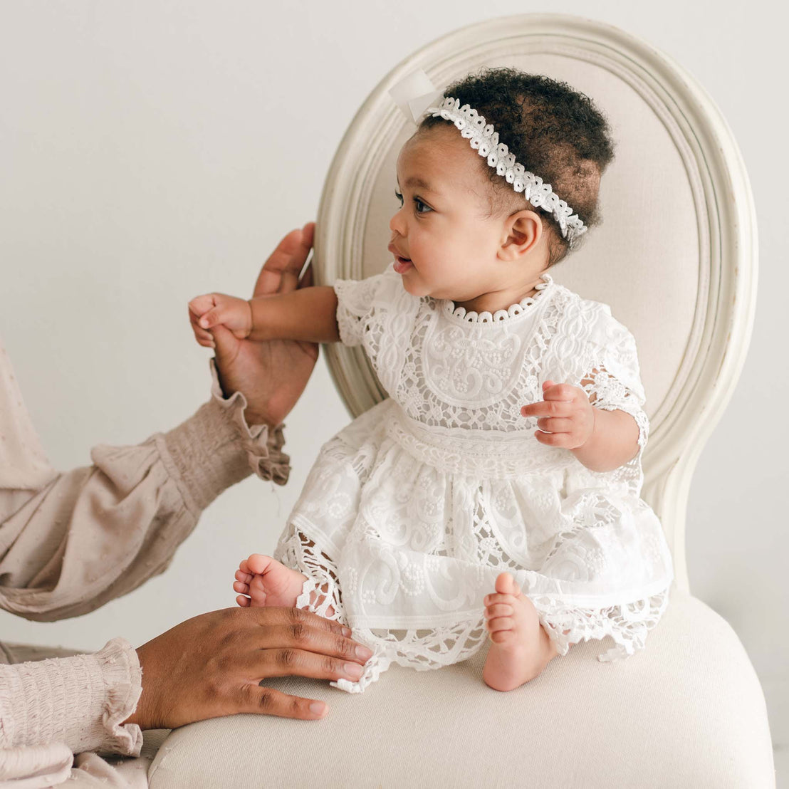 A baby wearing the Adeline Lace Dress & Bloomers with a matching headband sits on a beige chair. The child looks to the side while reaching out with one hand, supported partially by an adult from the side. The background is plain and light-colored.