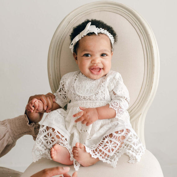 A cheerful baby wearing the Adeline Lace Dress & Bloomers and a coordinating headband sits on a cream-colored chair. The baby is grinning and grasping the hands of an adult who is partially visible, dressed in a beige long-sleeved top. The background is white and minimalist.