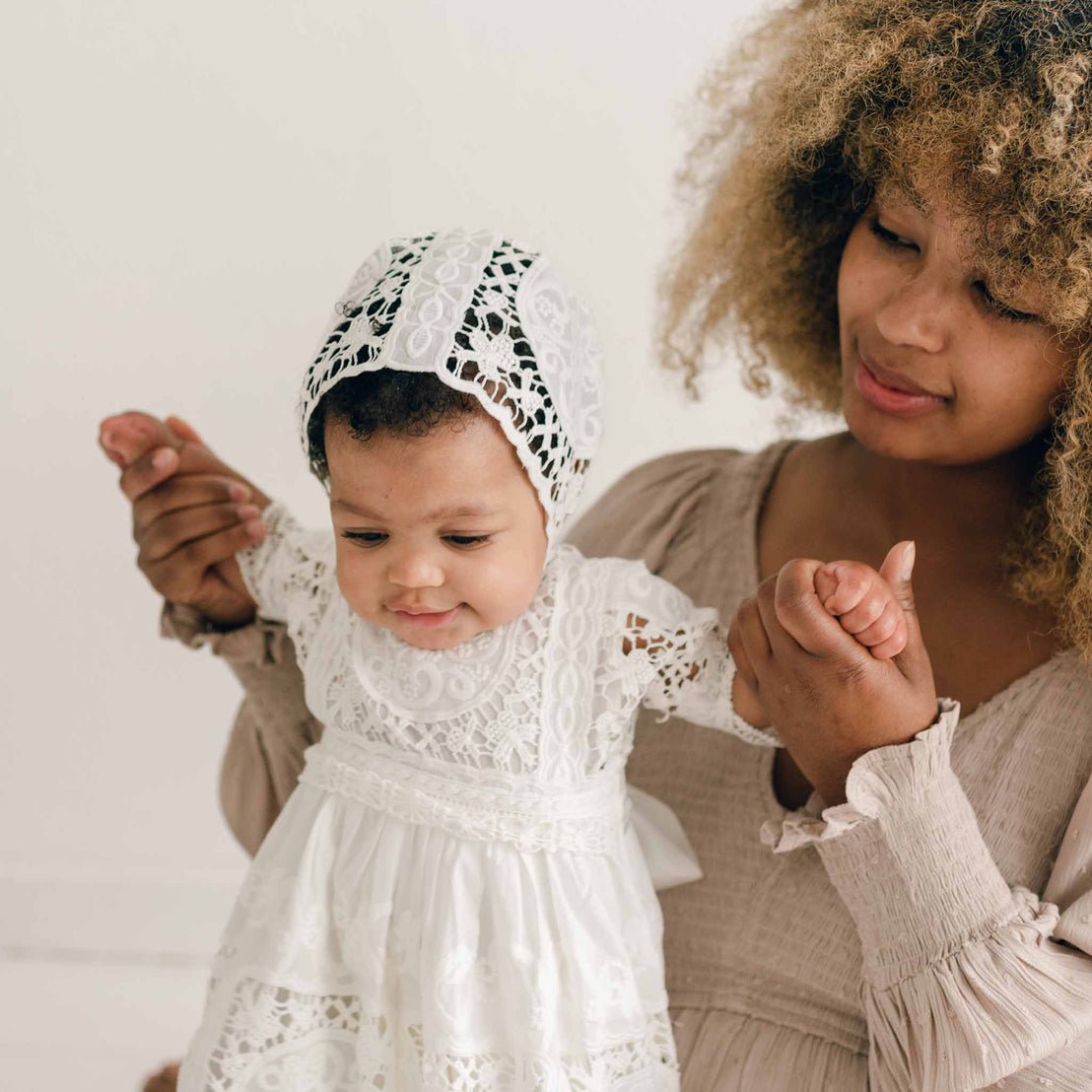 A woman with curly hair, wearing a beige blouse, gently holds the hands of a baby dressed in the Adeline Lace Dress & Bloomers with a matching bonnet. The baby looks down with a curious expression. The plain white background emphasizes their connection.