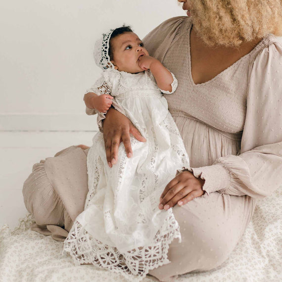 A woman with curly hair and a light-colored dress sits on the floor, holding a baby wearing the Adeline Lace Christening Gown & Bonnet, adorned with delicate cotton lace. The baby has a lace headband and is sucking on their hand while looking up at the woman.