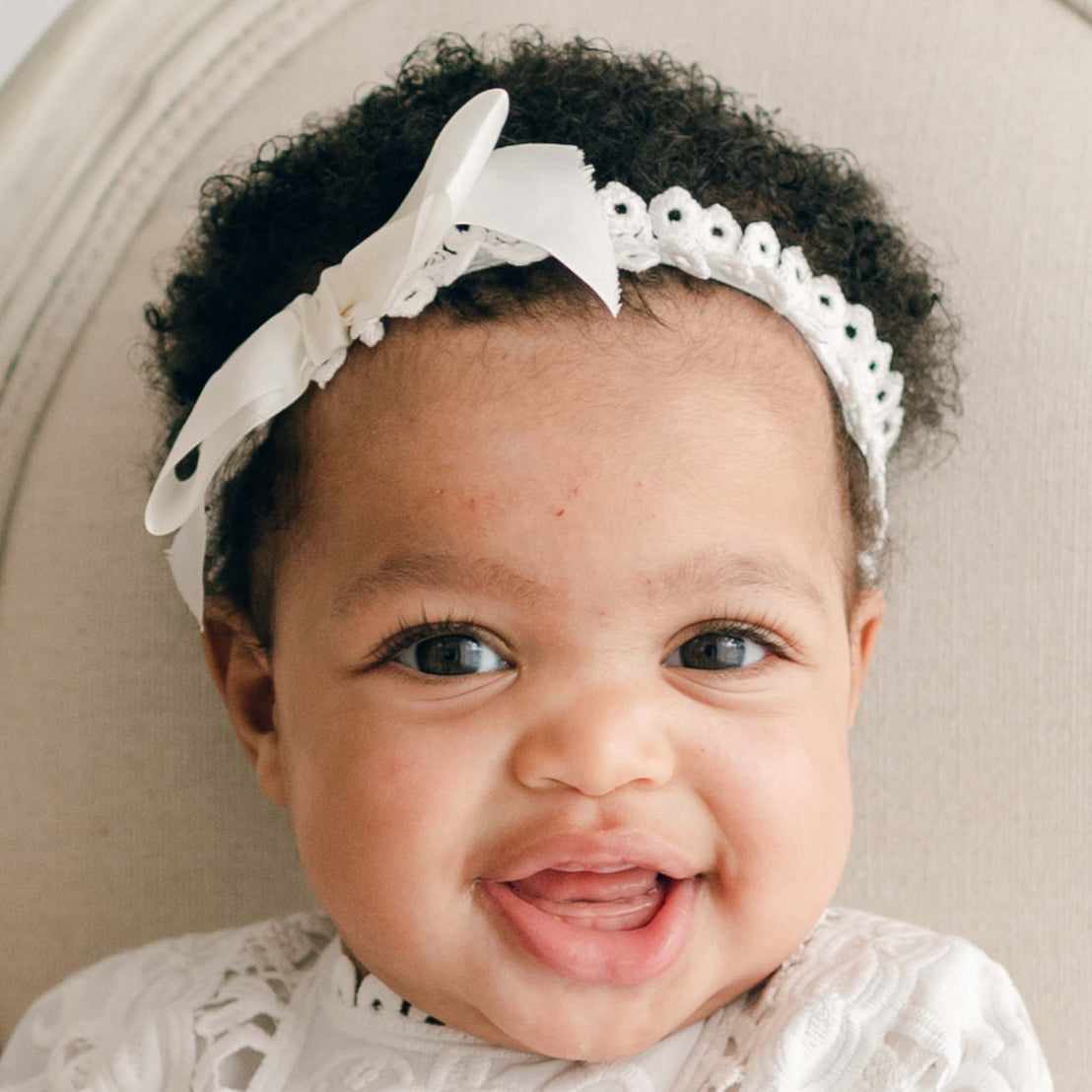 A smiling baby with curly hair wears the Adeline Headband, a white, lace-trimmed accessory featuring a silk ribbon bow. The baby is dressed in a white christening outfit and is seated against a light-colored cushion. The close-up captures the baby's cheerful expression and wide eyes.