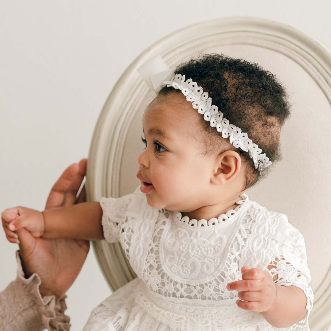 A baby wearing a white lace dress and an Adeline Headband with a silk ribbon bow sits in a round-backed chair. An adult's hand is gently supporting the baby from the side. The baby looks to the right with a curious expression.