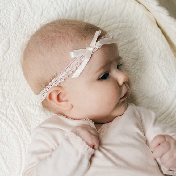 Close up of a newborn baby on a white quilt wearing an Ava Lace Headband with matching Ava Layette Coming Home Outfit. Details show blush silk ribbon bow with matching stretch lace.