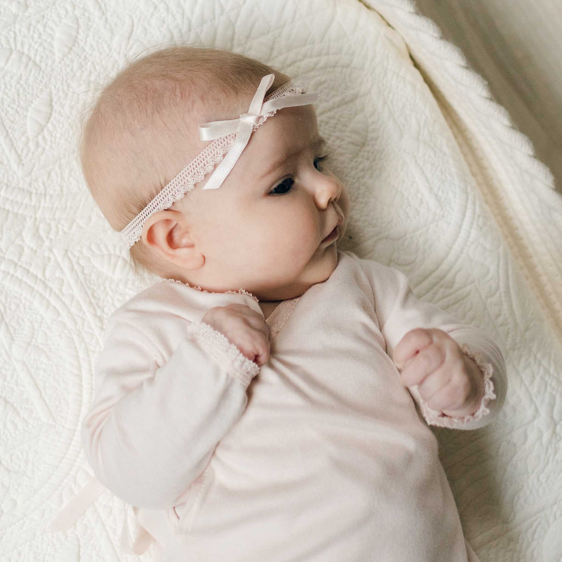 Close up of a newborn lying on a beige textured blanket in a crib. She is dressed in the Ava Layette coming home outfit for baby with matching blush ribbon bow headband. Part of the Baby Beau & Belle Baby Girl Coming Home Outfit Collection.