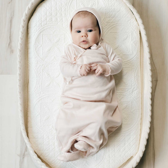 Newborn baby dressed in the blush pink cotton Ava Layette baby girl coming home outfit with quilt bonnet, and lying in a beige crib.