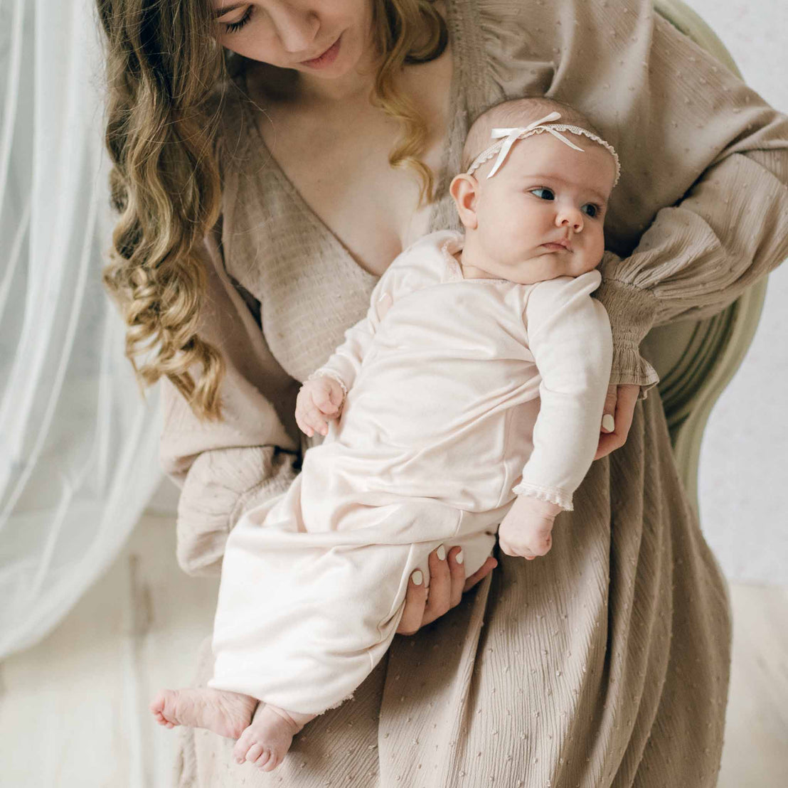 Newborn girl dressed in the Ava Layette coming home outfit and matching blush ribbon bow headband held gently by a woman in a beige dress. Set against a light backdrop with a sheer curtain.