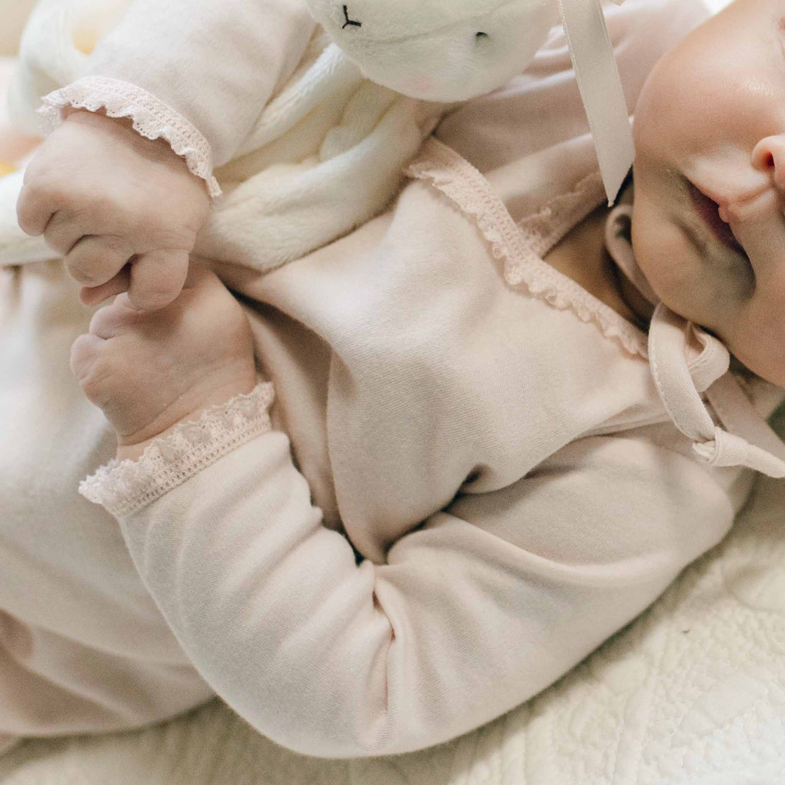 Close-up of a new baby in the Ava Layette baby girl coming home outfit showcasing the blush lace trim along the necks, cuffs and hem. The newborn holds a plush bunny toy in her right arm.