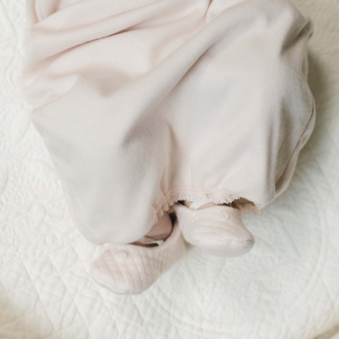Close-up of a newborn's feet in the blush pink Ava Quilted Booties peeking out from the skirt of the matching Ava Cotton Layette Coming Home Outfit. 