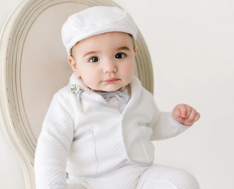 A baby wearing a white outfit and cap sits on a cream-colored chair. The baby looks directly at the camera with a neutral expression. The background is plain and light-colored.