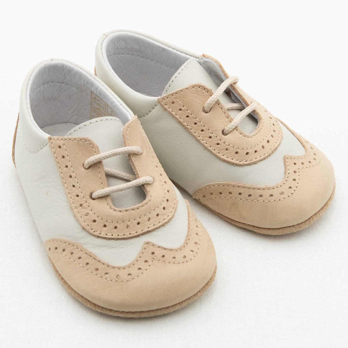 A pair of Beige & Ivory Wingtip Shoes made from beige and ivory leather rests on a white surface. The handmade leather shoes feature lace-up fronts and decorative perforations in the beige sections, adding a stylish, vintage touch. The soles are soft and designed for infants.