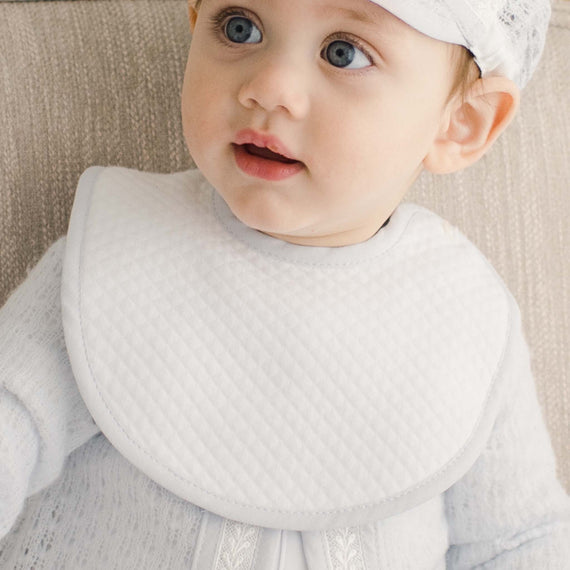 Baby boy wearing the Harrison Bib made with a very soft textured white cotton and features blue linen binding around the perimeter