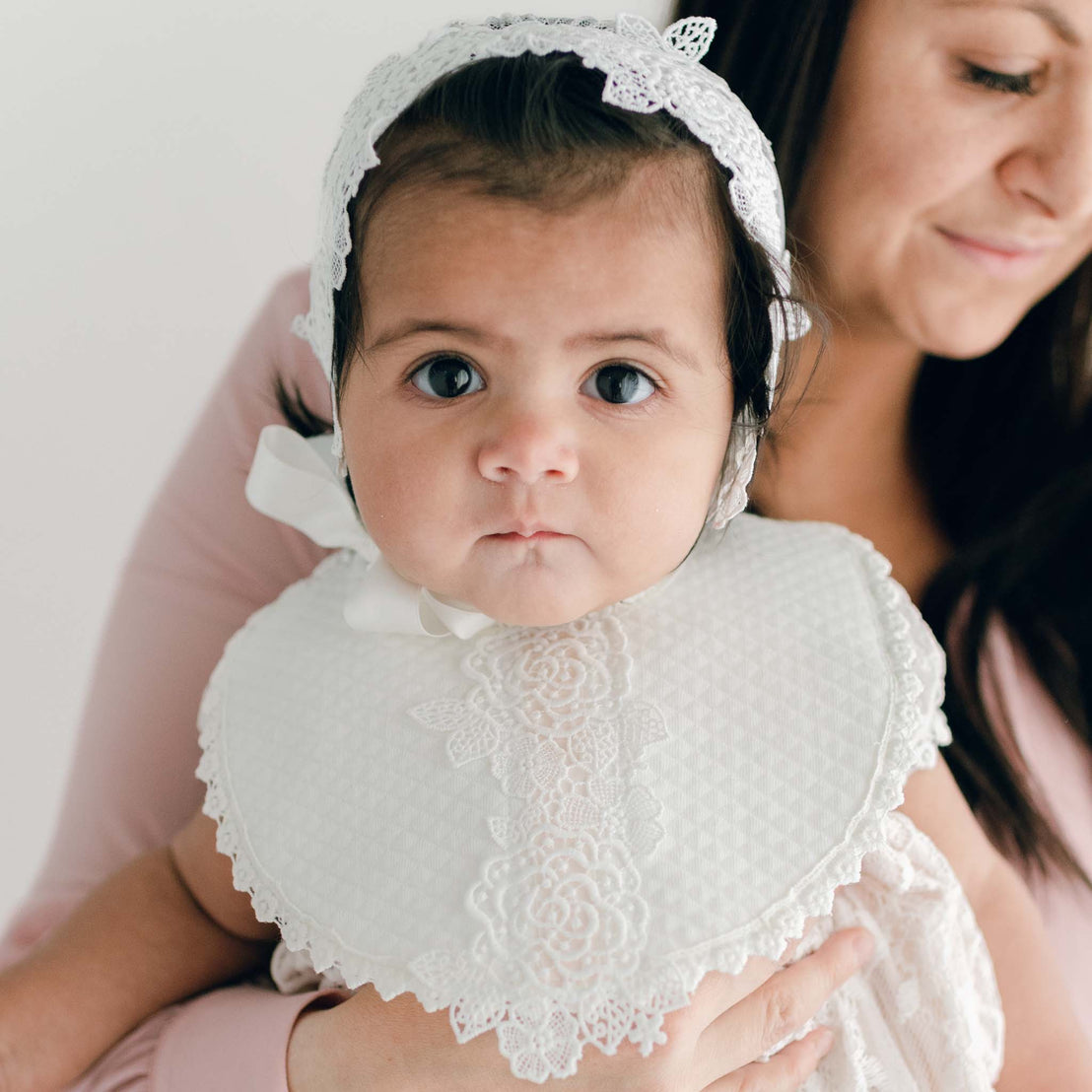 A woman holding a baby girl wearing a Juliette Bib and handcrafted white lace bonnet, both looking directly at the camera with a soft white background.
