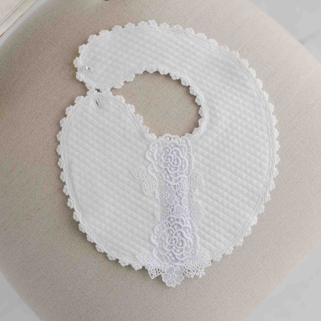 A white lace bib for babies, perfect for christening or baptism, featuring intricate floral patterns and scalloped edges, displayed on a light beige surface.