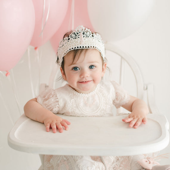 A toddler wearing a lace dress and the First Birthday Crown sits in a white high chair. Pink and white balloons are tied to the chair. She looks at the camera with a smile, her hand resting on the high chair tray in front of her.