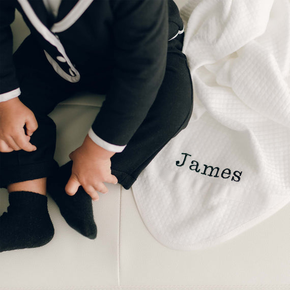 Baby boy with James Personalized Blanket. It is made from 100% white textured cotton with white grosgrain ribbon, silk ribbon, and the name "James" embroidered on the corner.