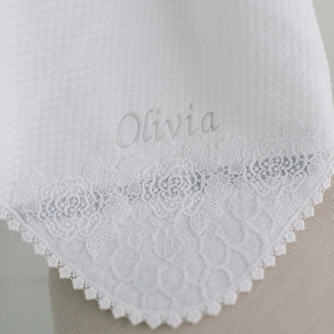 Close-up of a boutique white Olivia Blanket with delicate lace detailing along the edge and the name "Olivia" embroidered in a graceful, italic script.