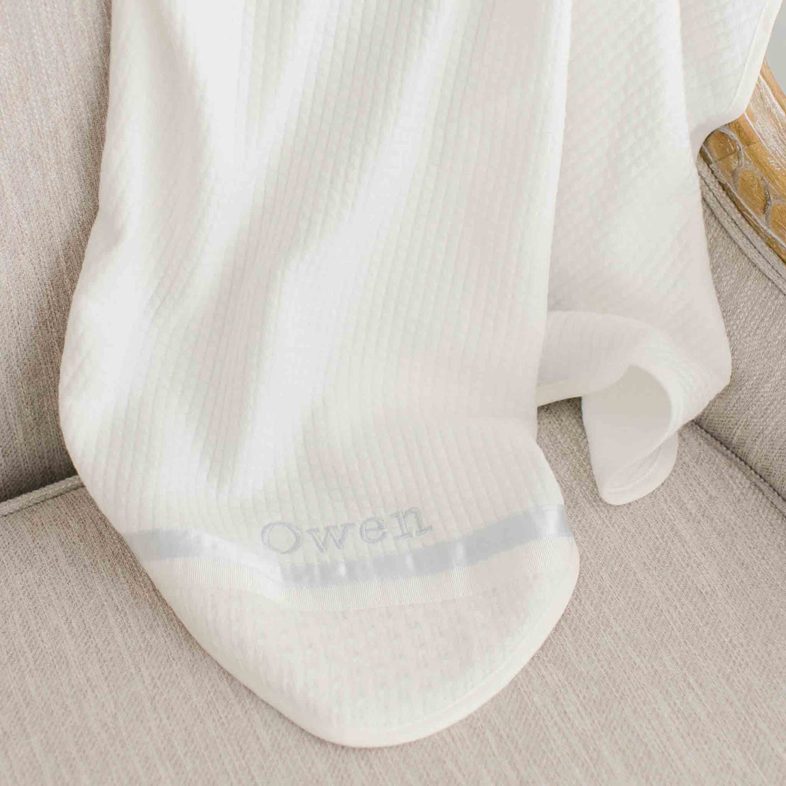 Photo of the corner of the Owen Personalized Blanket. The corner features an ivory knit and the name "Owen" embroidered with light blue thread