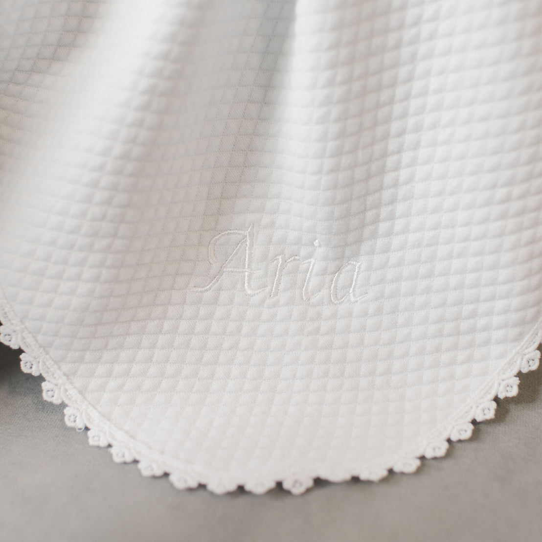Close-up of a white baby blanket with the product name "Aria" embroidered in white, featuring a small, delicate lace trim along the edge and perfect as a Baptism accessory.