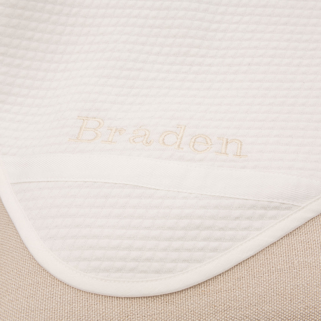 Flat lay photo of the Braden Personalized Blanket made from ivory textured cotton with an ivory grosgrain ribbon woven on the corner. The name "Braden" is embroidered on the corner of the blanket.