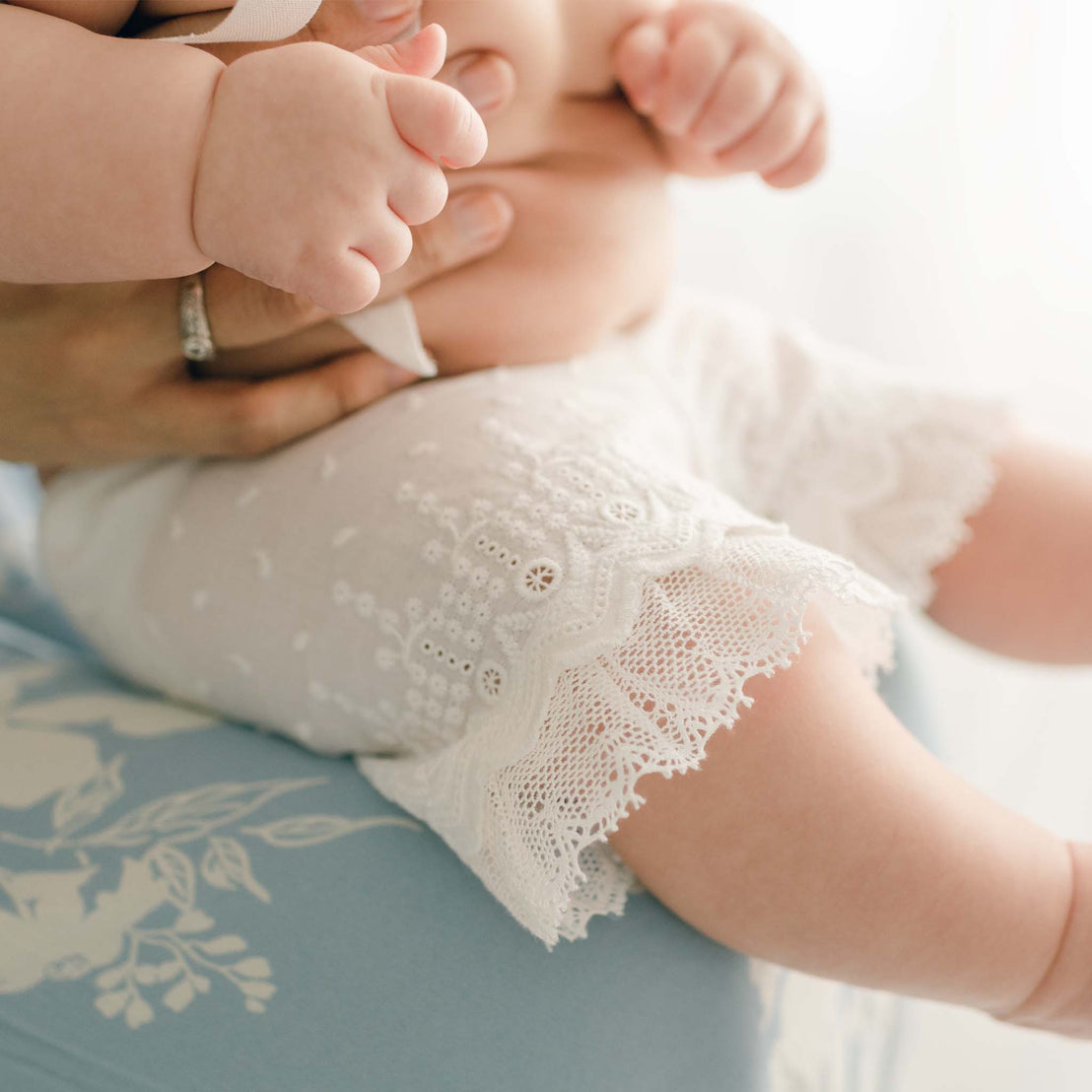 A close-up of a baby dressed in Emily Dress & Bloomers with a Green Sash for a christening, focusing on the baby’s legs and feet, against a soft blue and white vintage background.