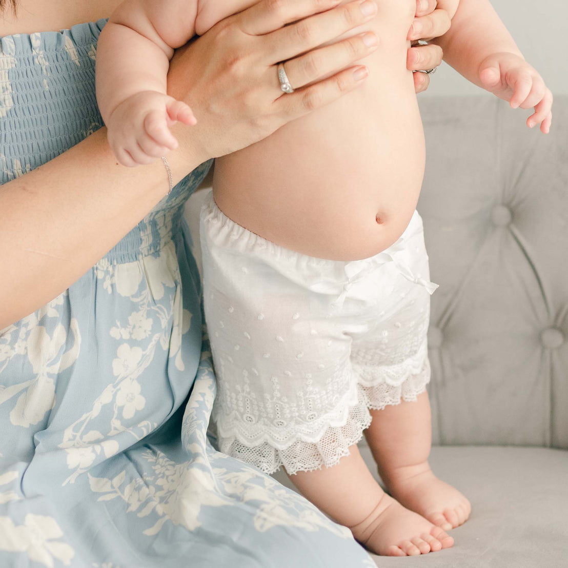 A mother in the Emily Dress & Bloomers | Green Sash holding her baby, dressed in white bloomers for a christening, standing on a bed with a soft gray background. Only the lower half of the baby's body