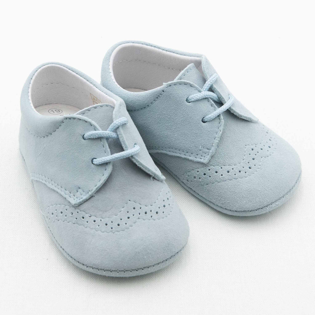 A pair of light blue baby shoes from the Baby Beau & Belle collection, known as Boys Suede Shoes, featuring lace-up closures and decorative perforations on the toe and sides to resemble a classic brogue design. These handmade suede baby shoes are elegantly displayed against a plain white background.