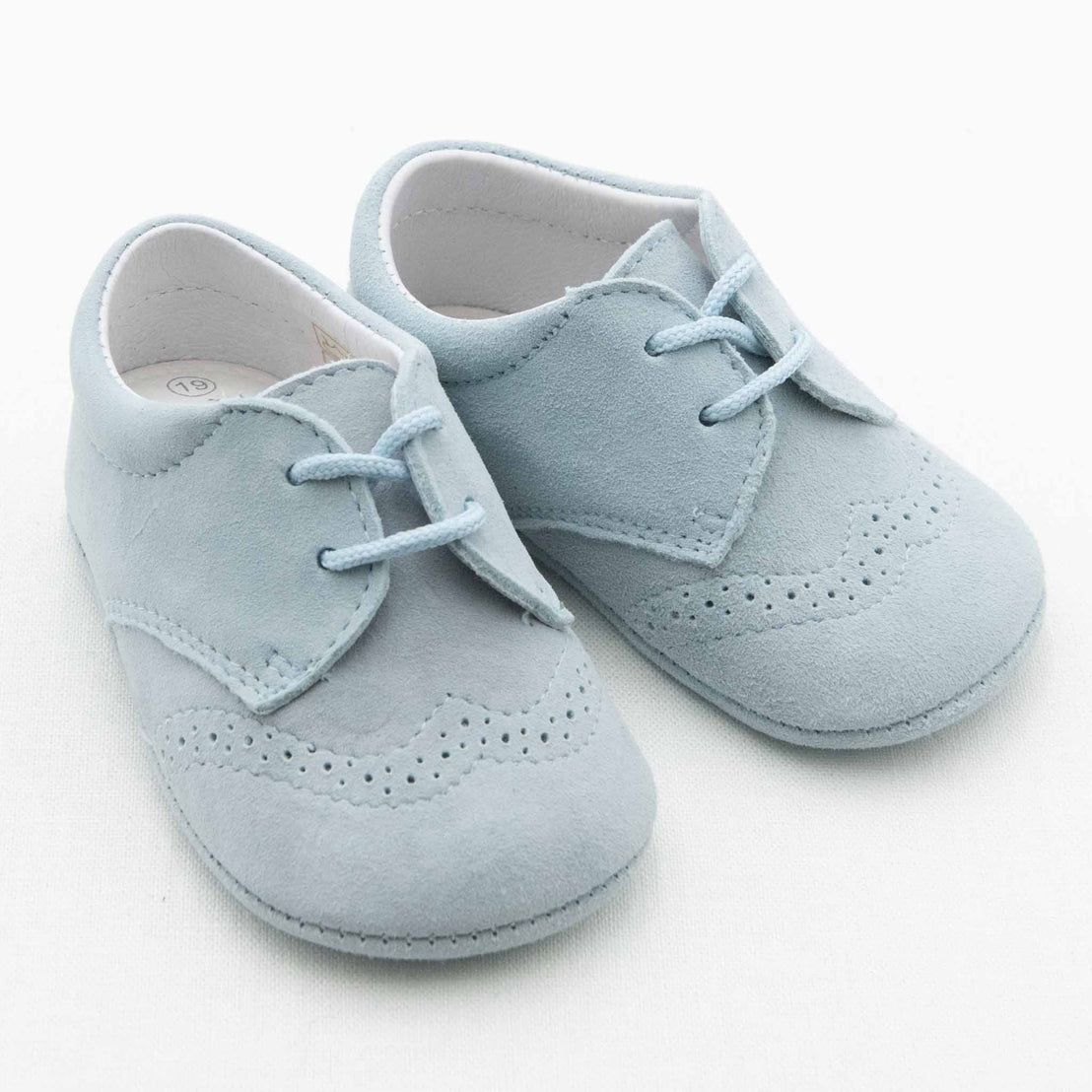A pair of Ian Suede Shoes in baby blue for boys, featuring detailed stitching and perforated patterns on the front and sides, with laces. These handmade suede shoes for infants are displayed on a plain white background.