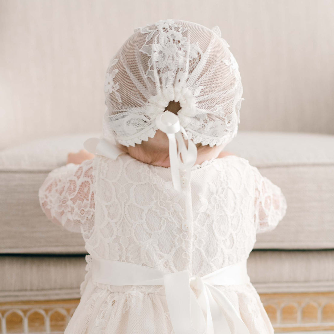 A baby dressed in an elegant white Juliette Lace Bonnet gown and bonnet, handcrafted with luxury boutique materials, standing with back to the camera.