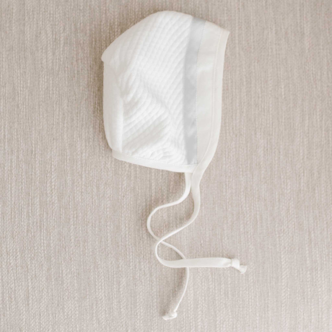 Flat lay photo of the Owen Quilted Newborn Bonnet made from ivory quilted cotton and a light ivory linen trim