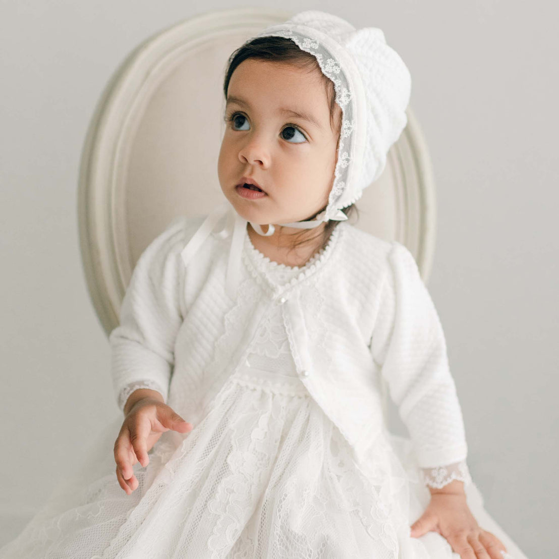 Baby girl sitting on a chair, wearing a quilted cotton sweater and the cotton baptism bonnet, part of the Victoria collection.