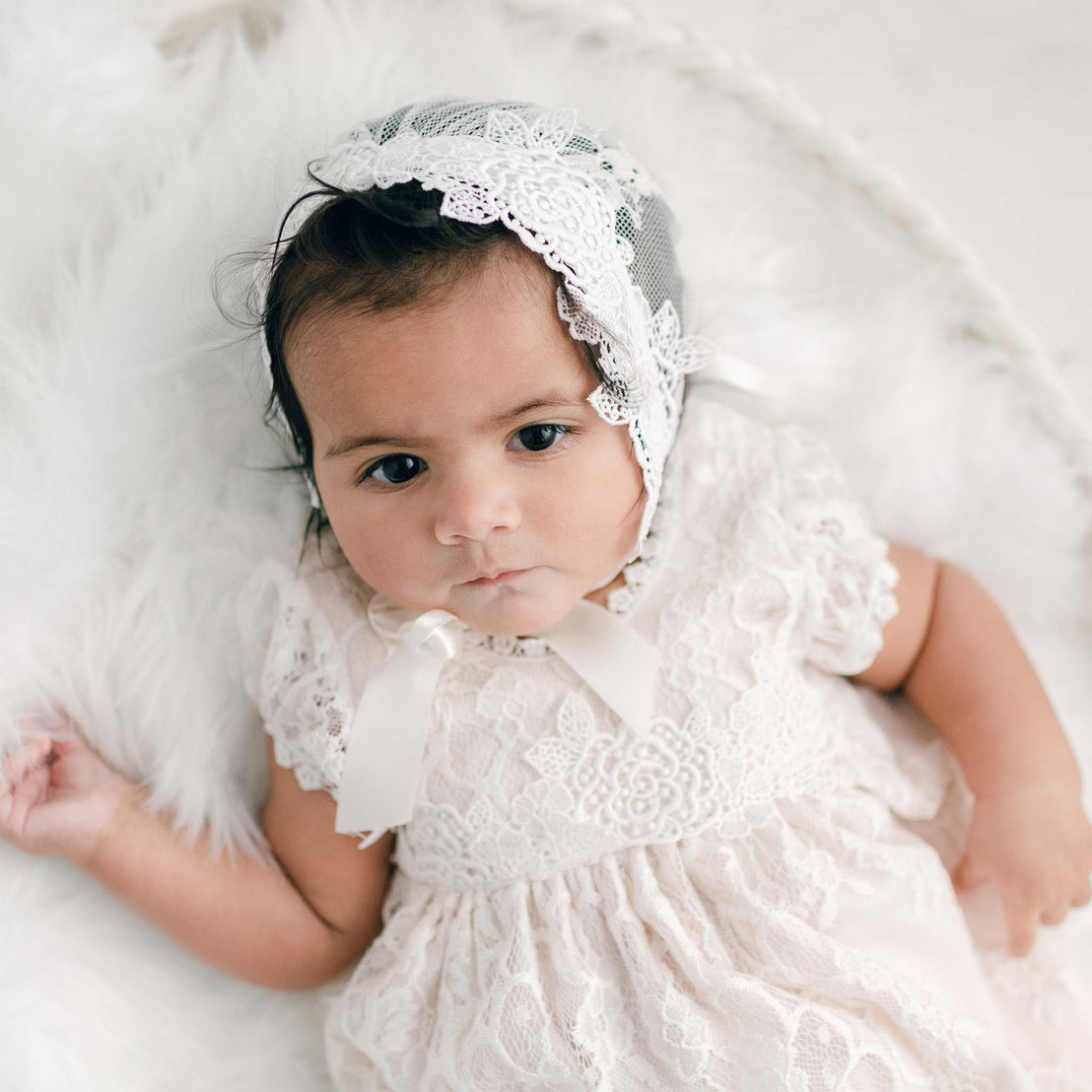 A baby with a thoughtful expression, wearing a vintage, handcrafted white lace dress and a Juliette Lace Bonnet, lies on a fluffy white blanket.