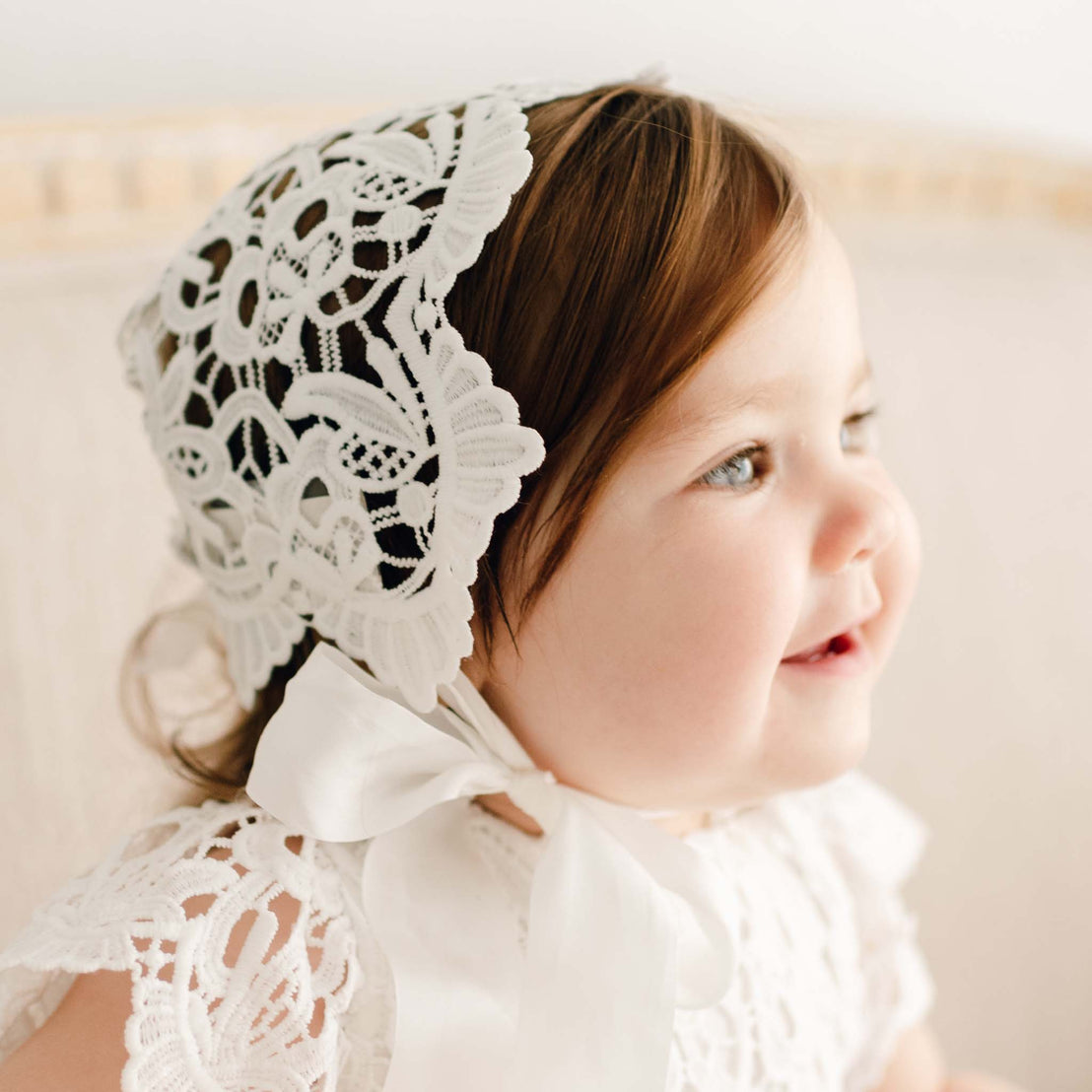 Close up of a baby girl wearing the Lola Lace Bonnet, uniquely designed with richly embroidered light ivory lace and cotton floral edge lace at the back. 