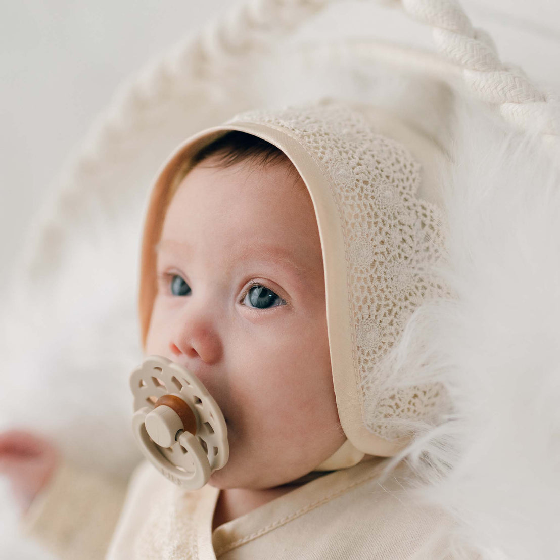 A close-up of a newborn girl with blue eyes and a pacifier, wearing a Mia Knot Gown & Bonnet, lying on a white fluffy blanket.