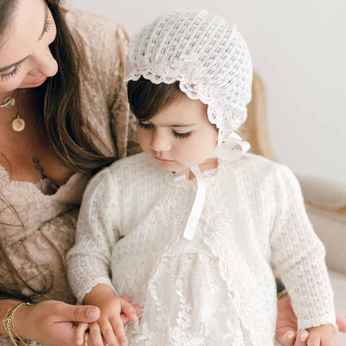 Baby girl wearing knit sweater and bonnet, holding her moms hand.