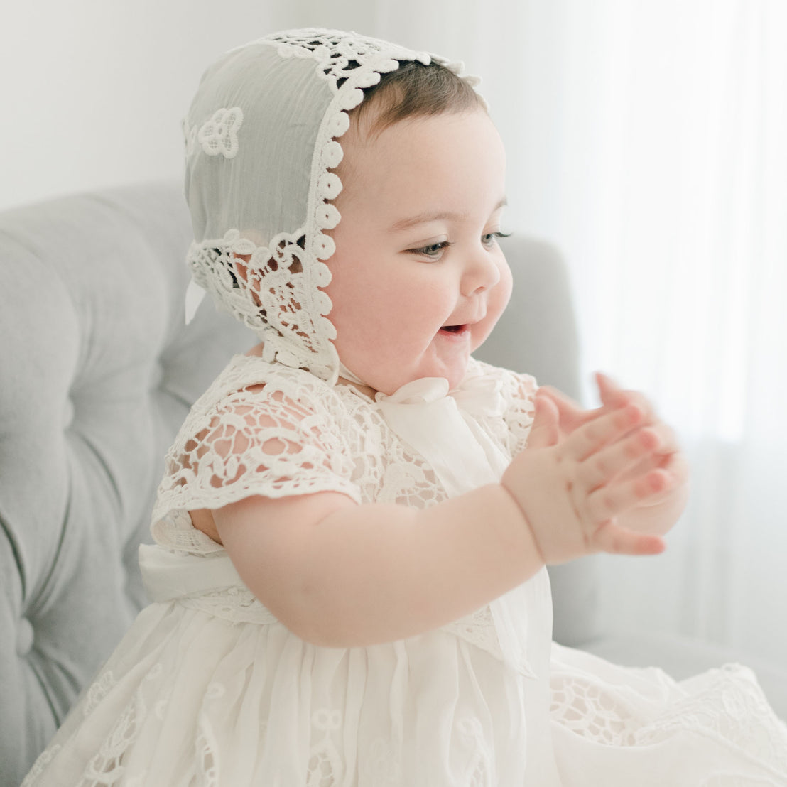 A baby claps happily, wearing the Grace Lace Bonnet paired with a delicate white dress featuring a silk ribbon. Seated on a gray chair, the softly lit background enhances the serene atmosphere of this christening day scene.