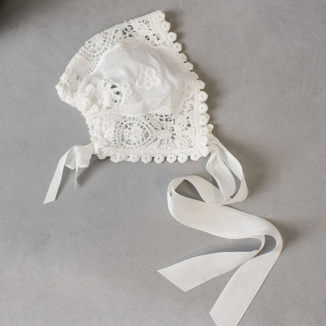 Introducing the Grace Lace Bonnet: a delicate ivory baby bonnet made from lace with intricate embroidery and scalloped edges. It features long satin ribbons and showcases detailed craftsmanship, displayed flat on a light gray surface.