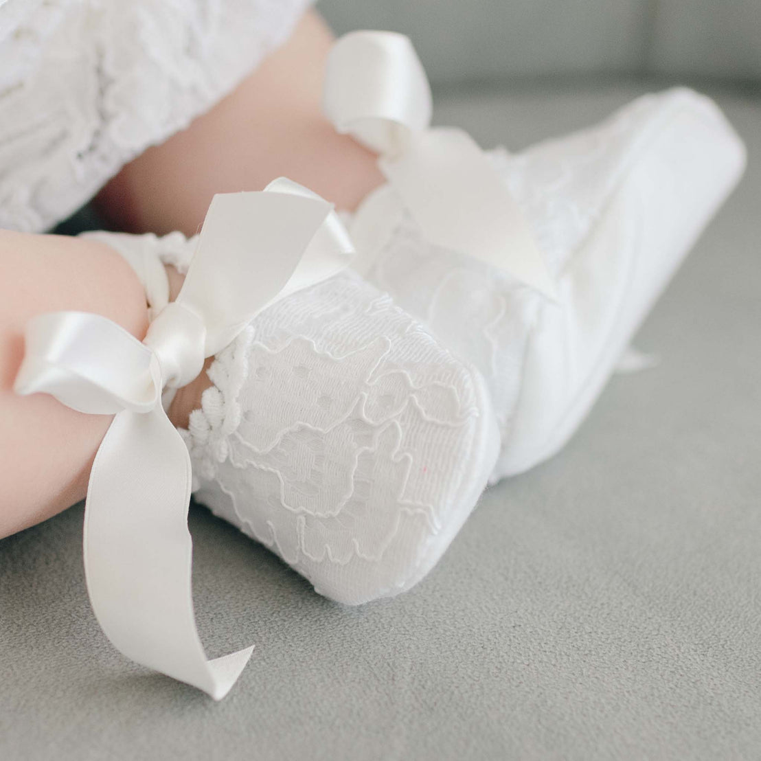 Baby girl wearing the Aria Lace Christening Booties made with white embroidered lace, lined with soft pima cotton, and fitted with white silk ribbon ties.