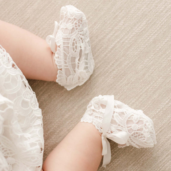 Baby wearing a pair of Lola Booties made with an all-over lace in a light ivory color and a silk bow tie.
